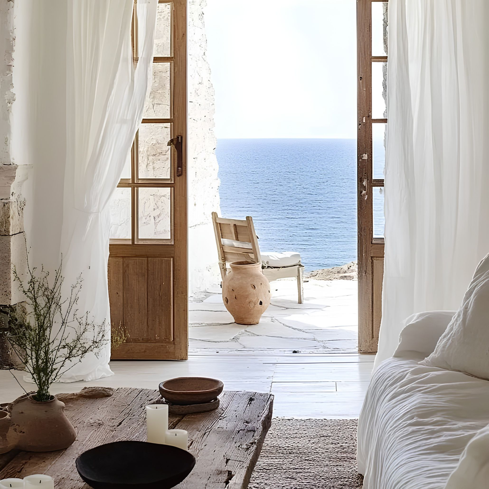 A whitewashed living space with billowing linen drapery, a weathered wooden door and a large terracotta vessel opening onto a cliffside sea view, capturing the quiet wood texture and craftsmanship of rustic greek and Mediterranean Interior design.