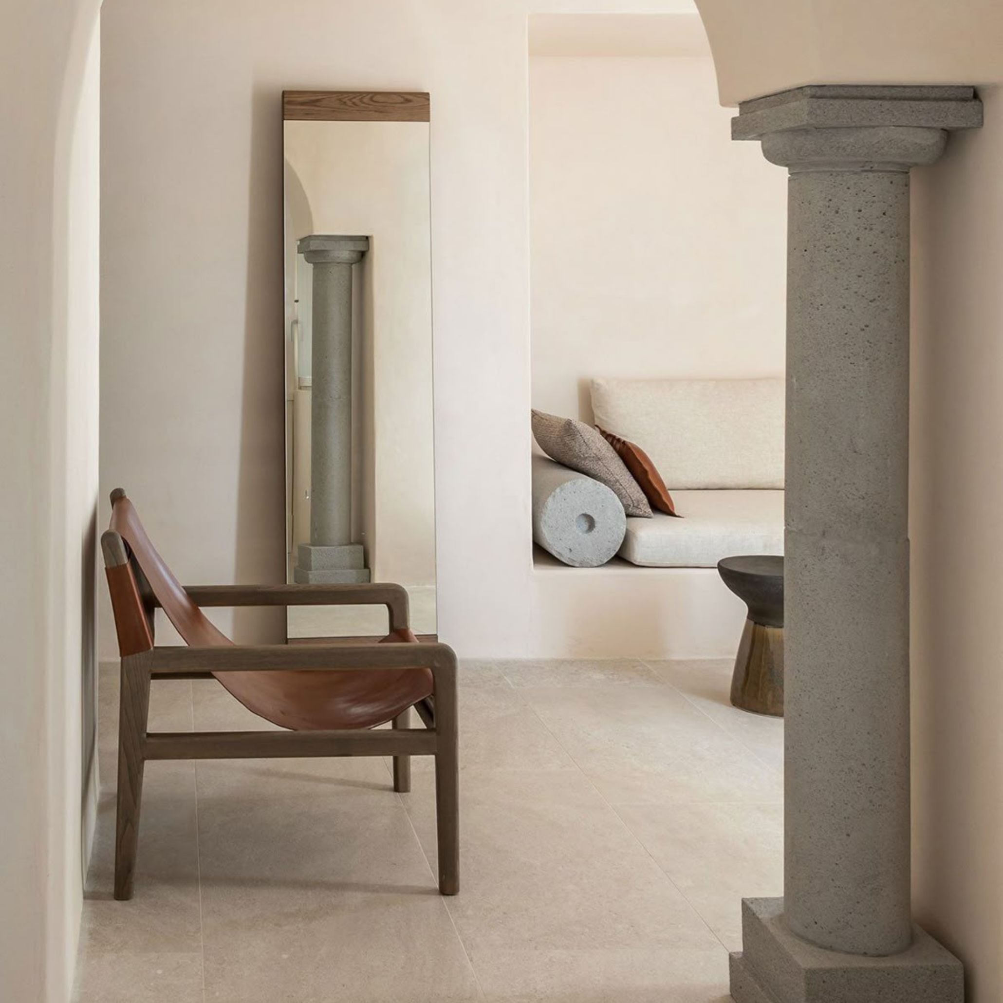 A pared-back hallway featuring a leather-and-oak lounge chair, a tall wall mirror and a carved stone column beside a built-in banquette, showing the mirror design and luxury living room furniture sensibility of rustic greek interiors.