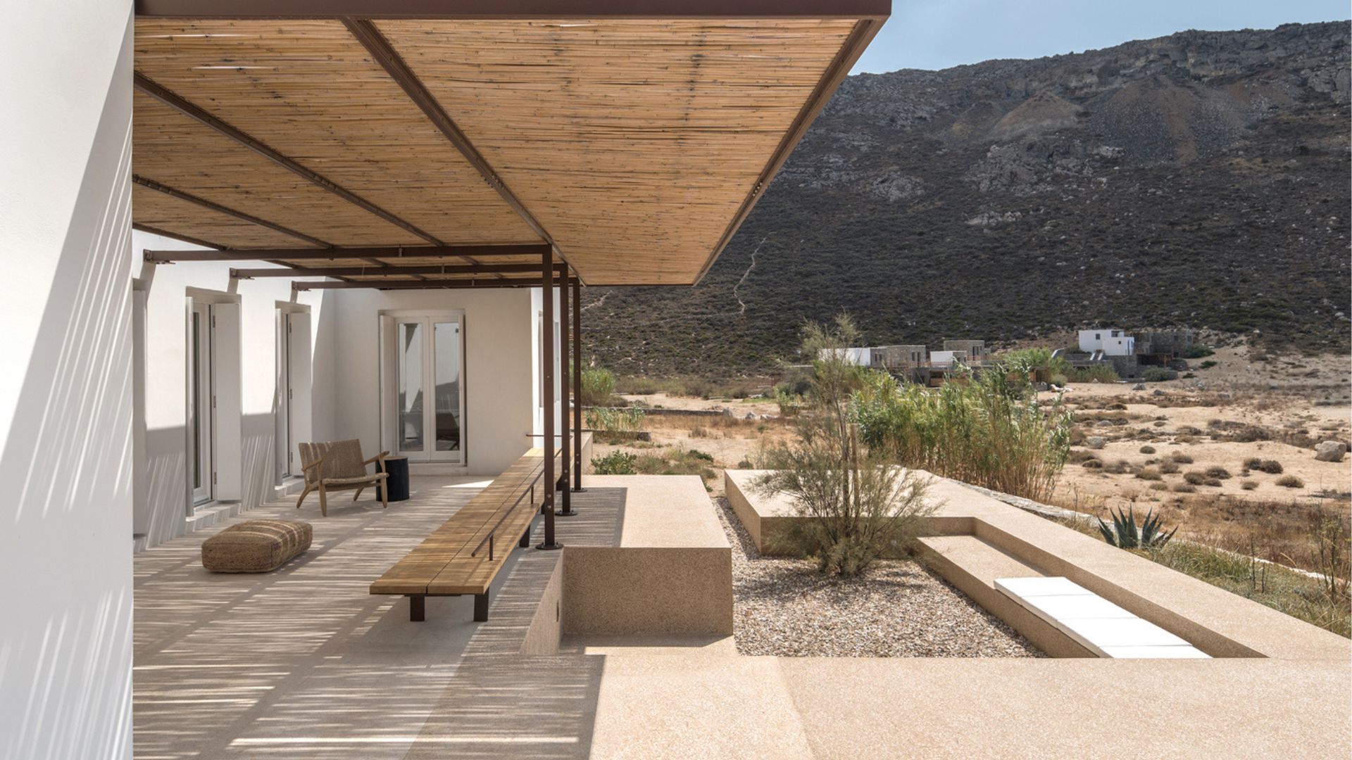 A shaded terrace with a reed-covered pergola, wooden bench and woven armchair overlooking arid mountain landscape, illustrating alfresco living and luxury home furniture true to rustic greek and Mediterranean Interior design.