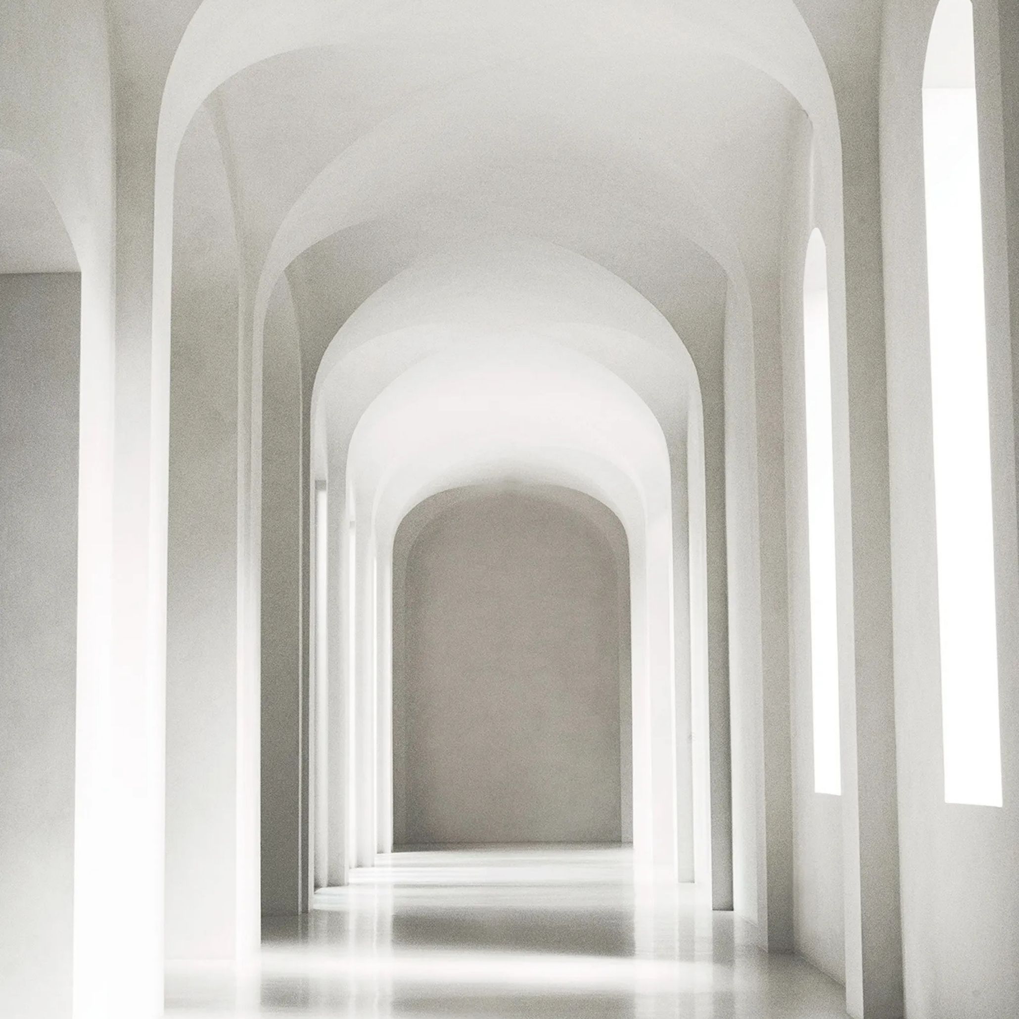A minimalist arched corridor in soft white tones, with a sequence of vaulted archways leading toward a sunlit end wall, evoking the calm, architectural quality found in living room architecture and pared-back interiors.