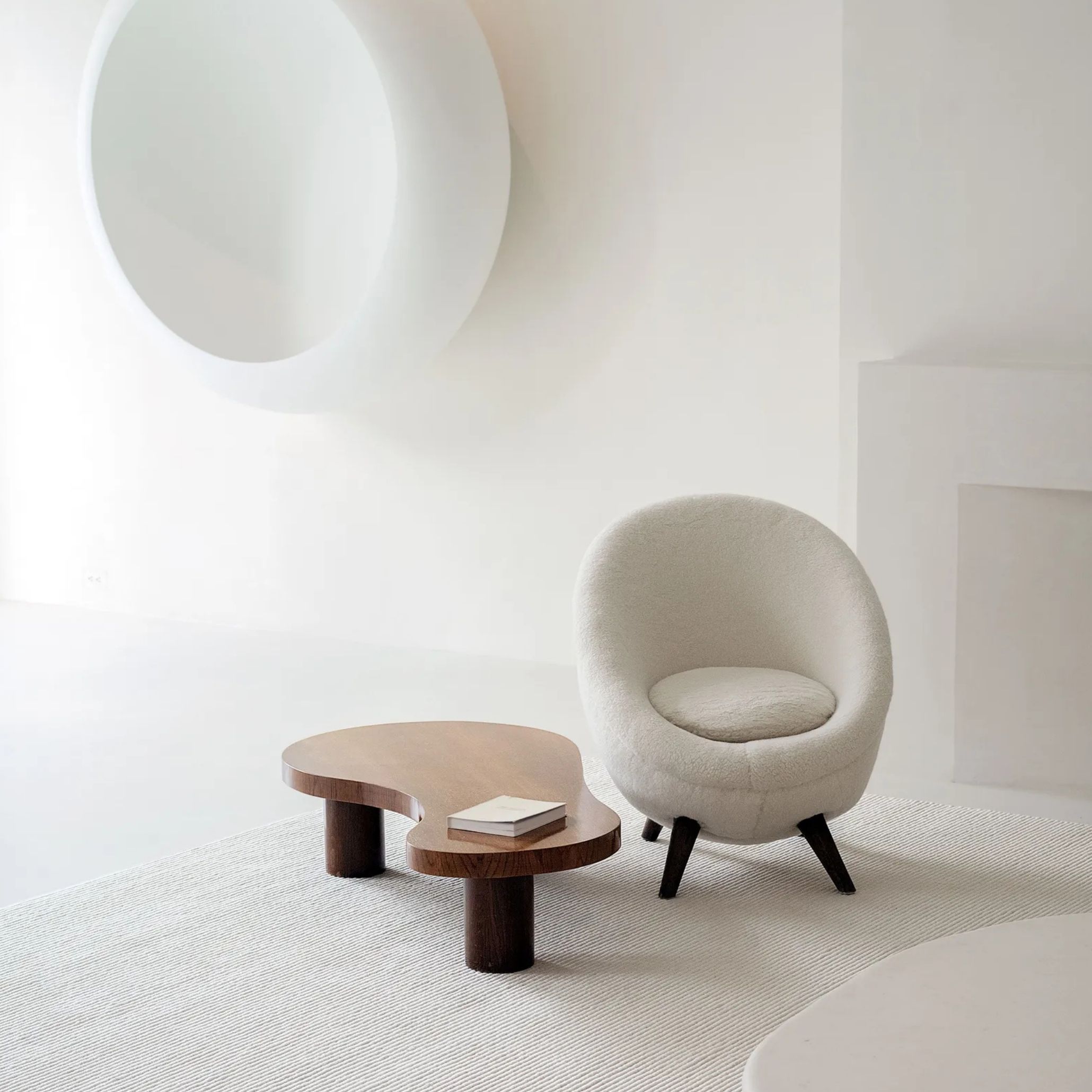 A serene living room corner featuring a rounded bouclé armchair in soft ivory beside an organic, curved wood coffee table with tapered cylindrical legs, set against plain white walls and a round mirror. The sculptural, restrained styling reflects the kind of luxury living room furniture and interior design inspiration associated with quiet, considered spaces.
