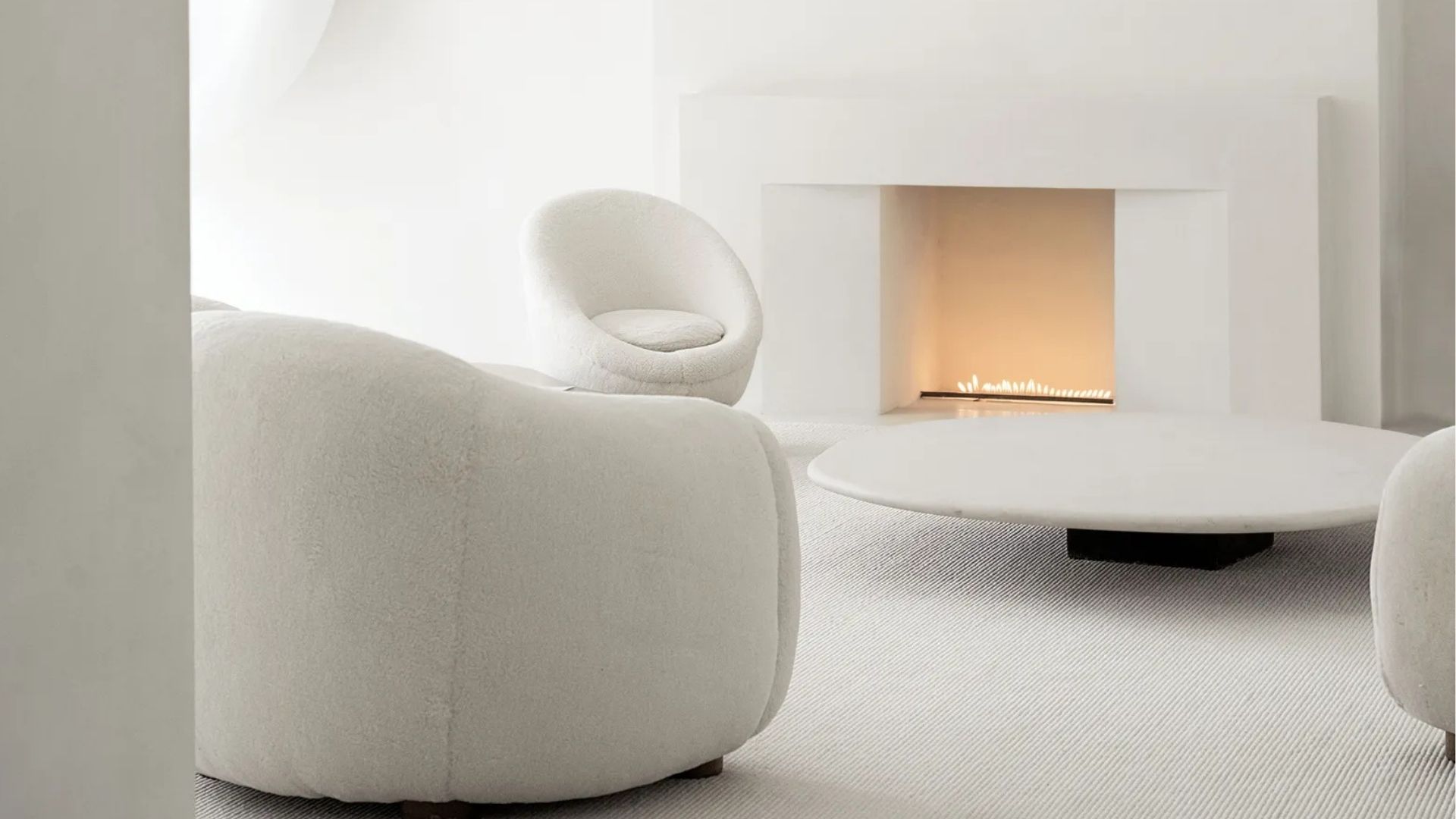 A minimalist living room in soft white tones, featuring an oversized bouclé sofa and a rounded armchair beside a sculptural white fireplace and a smooth, low round coffee table. Soft curves, tactile fabrics and a cohesive neutral palette create a sense of quiet luxury, reflecting the kind of restrained, proportion-led interior design inspiration behind Kim Kardashian's living room.