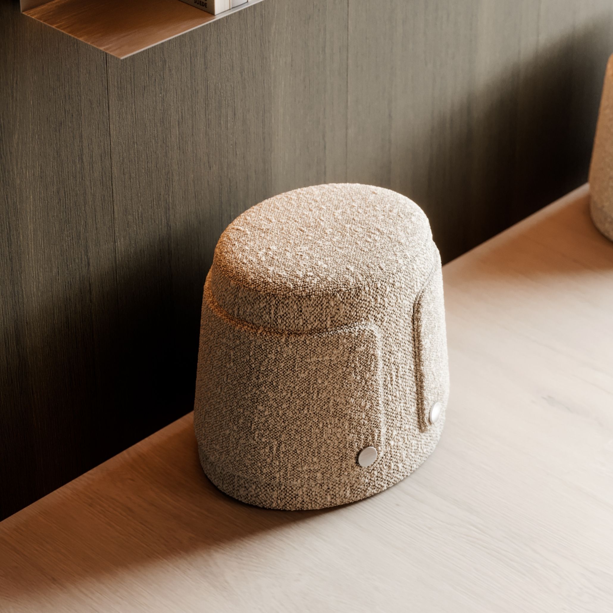 The Dolomites Stool in a warm bouclé, its rounded, barrel-like silhouette detailed with visible stitching and metal button fasteners, set on a wooden shelf against a dark wood-panelled wall. The tactile furniture upholstery and compact form reflect the sculptural, material-led character that defines this piece of interior design inspiration.