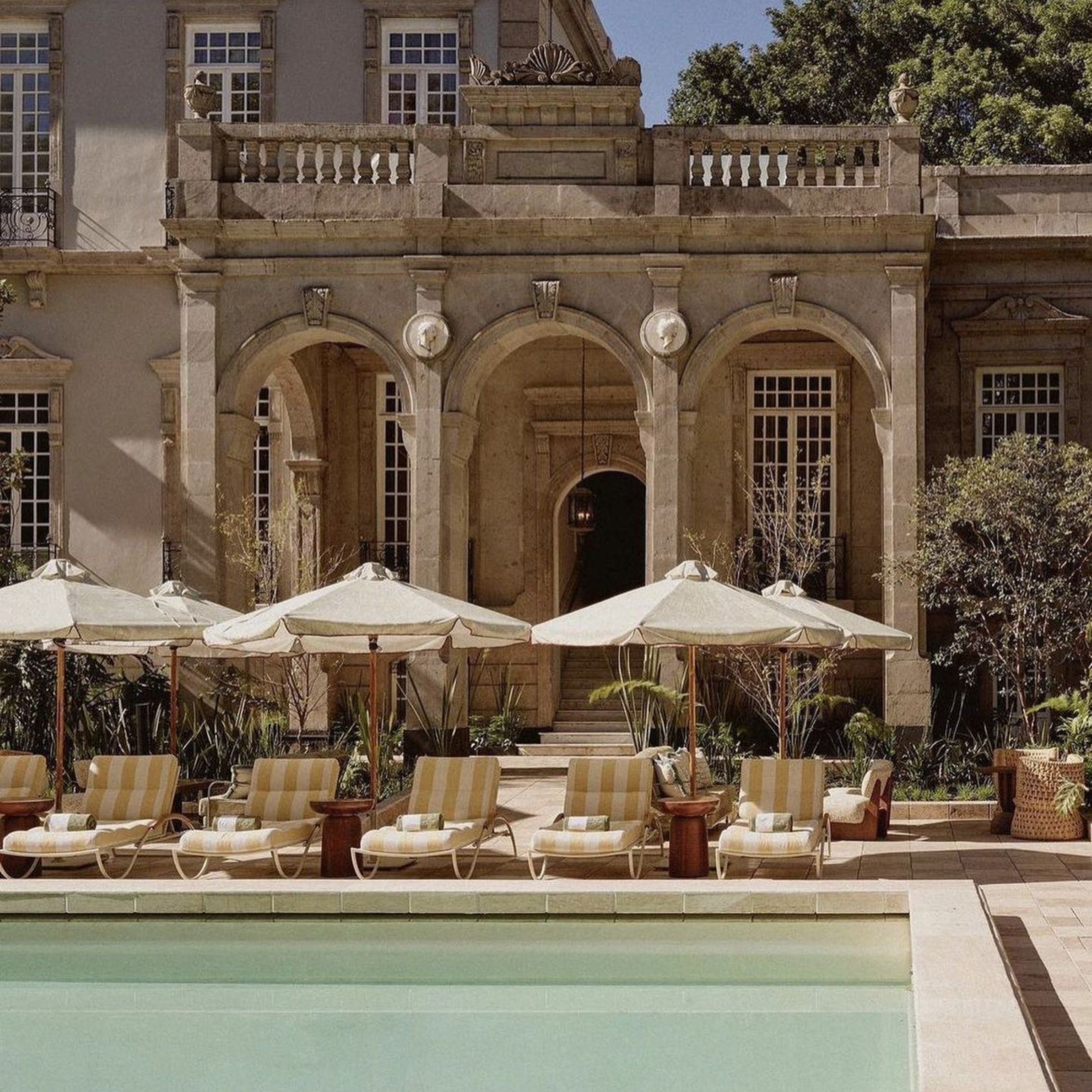 Striped mustard-and-cream lounge chairs line a mint-toned pool beneath a grand stone façade with arched entryways and canvas umbrellas, an example of luxury home furniture shaping modern coastal interior design.