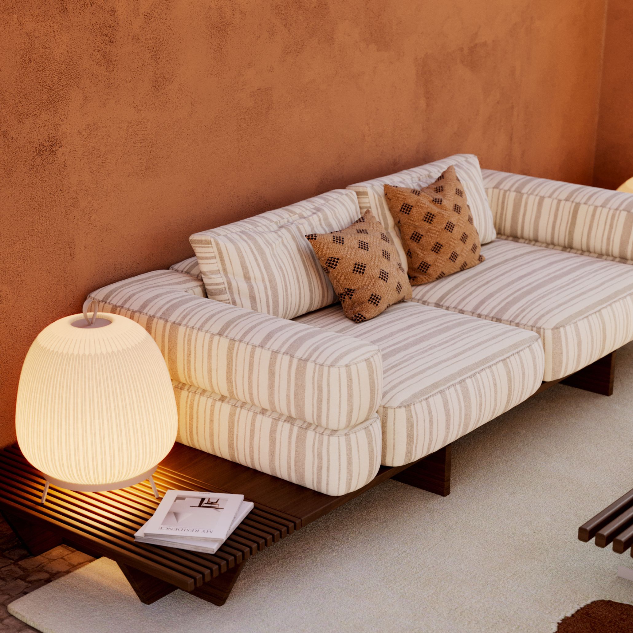 Detail of the Ripa Sofa in beige-and-cream striped outdoor furniture upholstery, styled with patterned throw pillows beside a slatted wood side table and glowing lantern lamp against a terracotta wall.