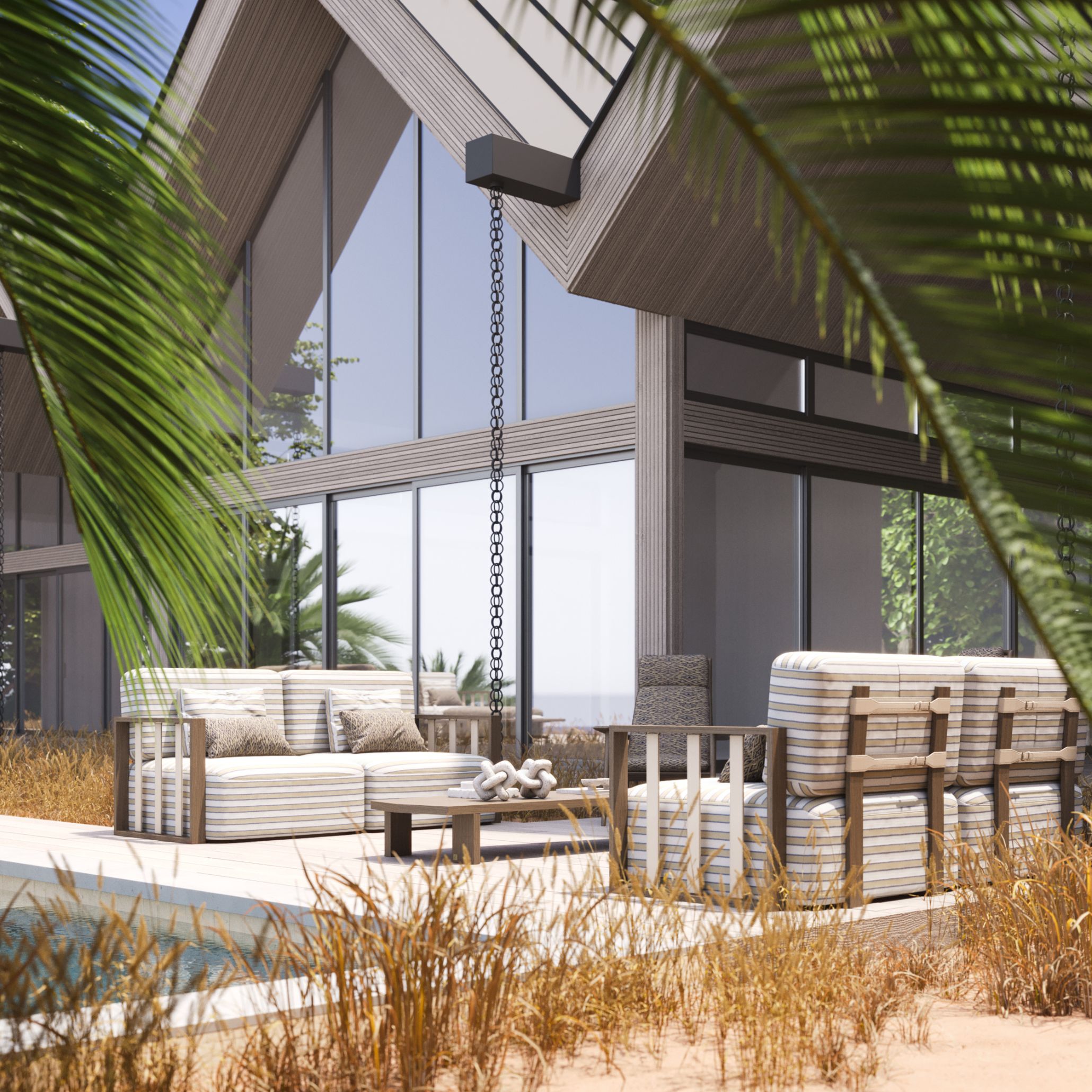 A poolside terrace featuring blue-and-white striped outdoor furniture sofas and armchairs beneath a slatted wood pergola, framed by dry grasses and palm fronds, an example of Riviera Lines brought outdoors.