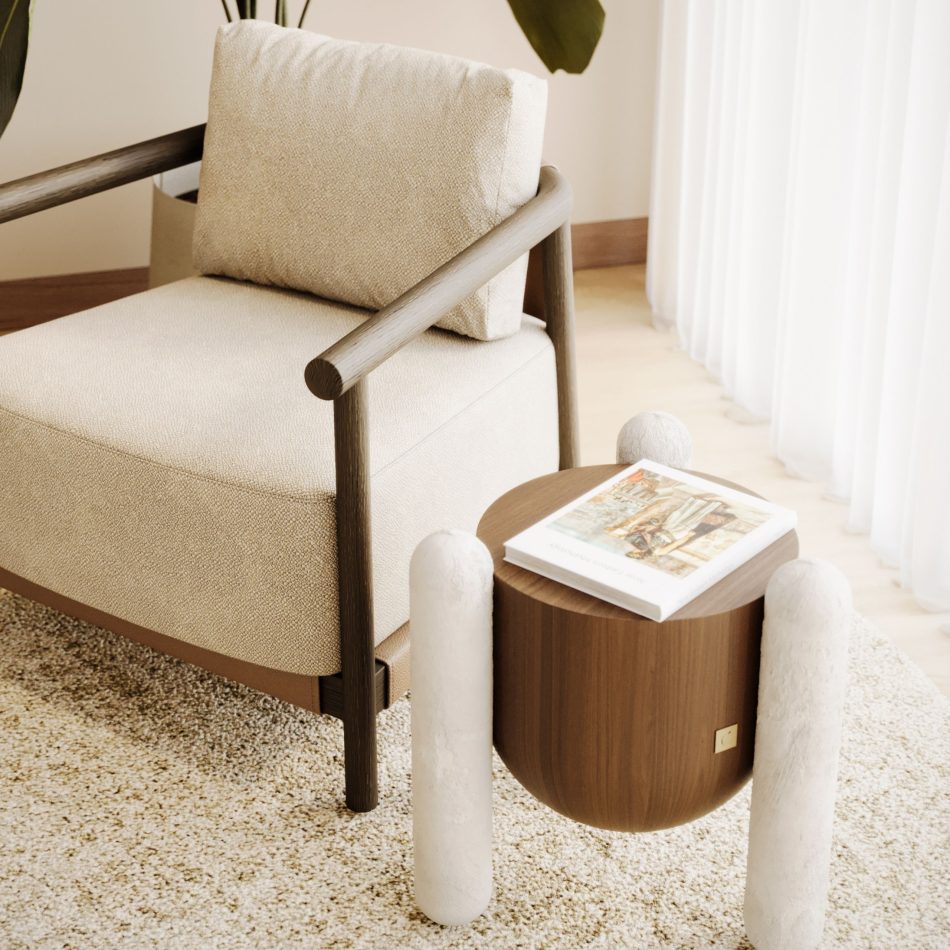 A close up of a living room corner, showing a beige upholstered armchair with a slender dark wood frame set against a neutral, sunlit interior. Beside it sits the Antónia side table, its rounded marble legs cradling a curved walnut body topped with a book, bringing a sculptural touch of modern design to the seating area. The pairing shows how this handmade furniture piece can sit comfortably within a warm, textured living room furniture setting.