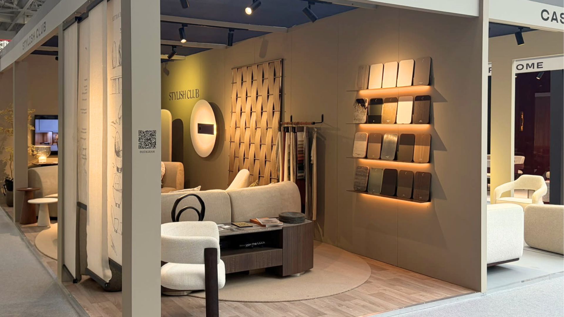 A warm, inviting corner view of Stylish Club's exhibition stand at Decorex 2026, featuring organic curved seating, a low coffee table, woven wood wall panels, and backlit material swatches. The booth explores forms and proportions that deliver timeless interior design inspiration, organic modern furniture, and custom furniture.