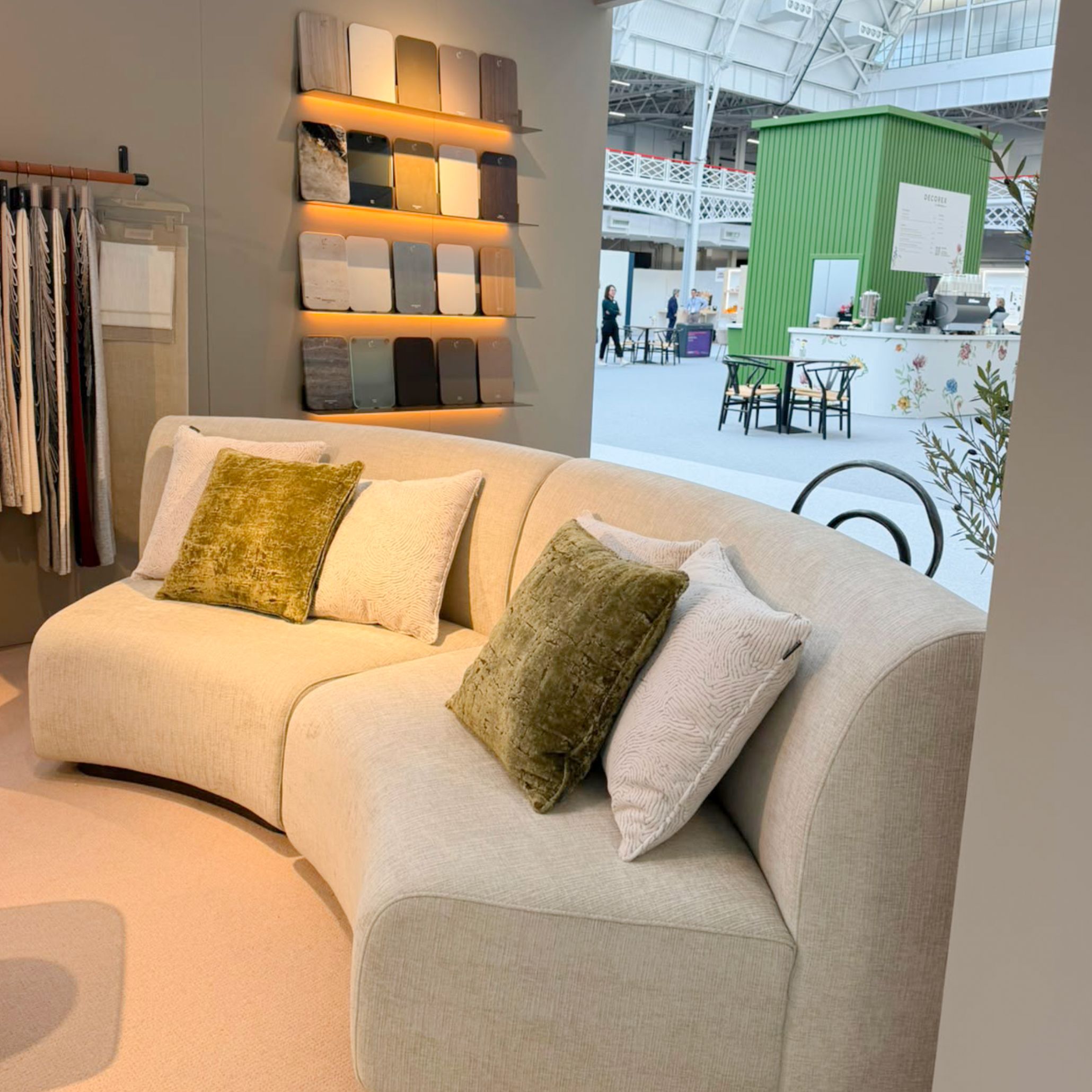 A close-up view of Stylish Club's Stone Sofa displayed at Decorex 2026, featuring river stone-inspired organic contours and plush textured upholstery accented with olive green throw pillows. Positioned near illuminated material swatches, this piece provides sophisticated living room inspiration, custom furniture, and high-end living room furniture.