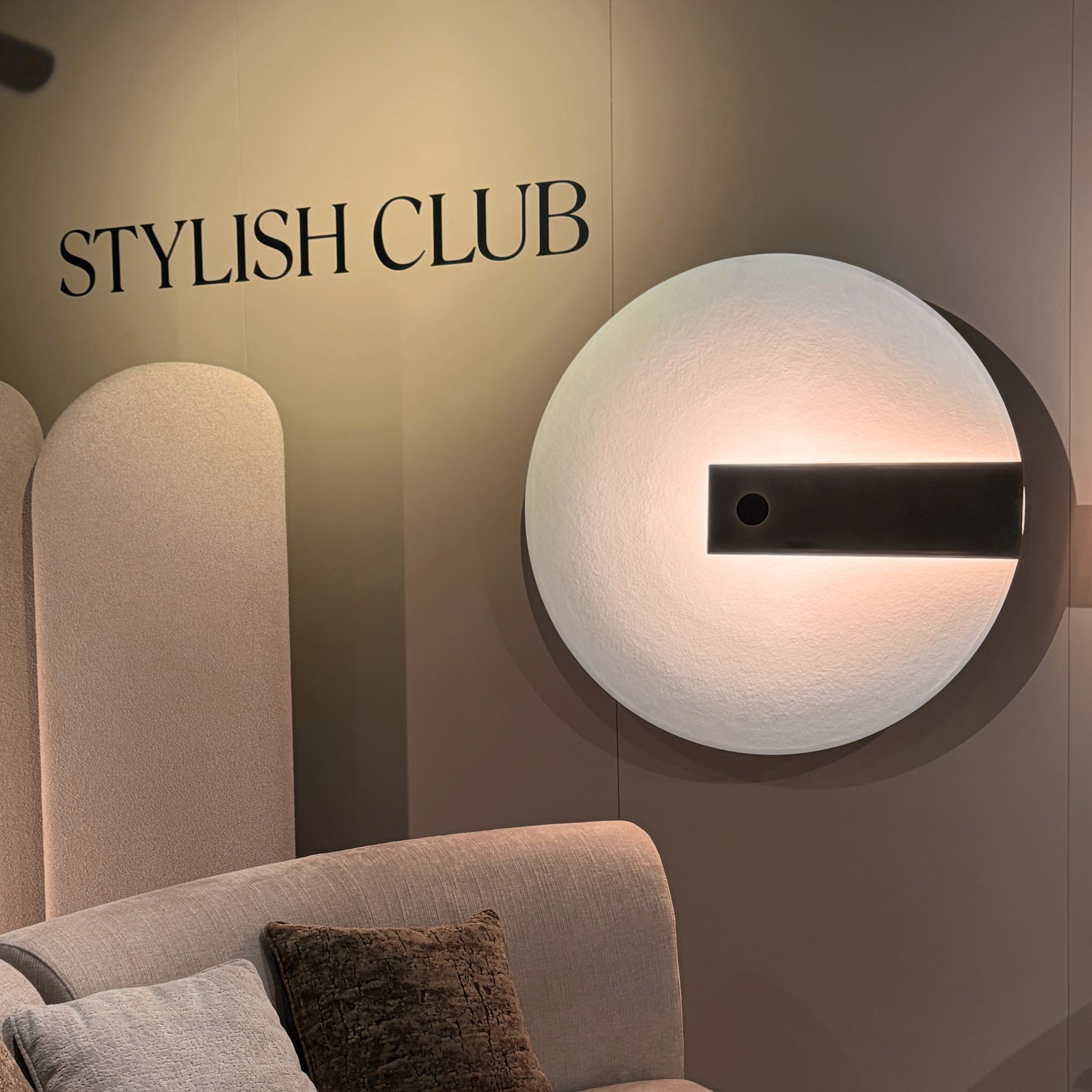 A detailed close-up of the circular Meridiana Wall light designed in collaboration with Hind Rabii Lighting Studio, mounted above plush textured seating at Stylish Club's Decorex 2026 booth. The warm backlight highlights delicate surface textures, adding sophistication to contemporary living room design, custom furniture, and overall furniture luxury design.