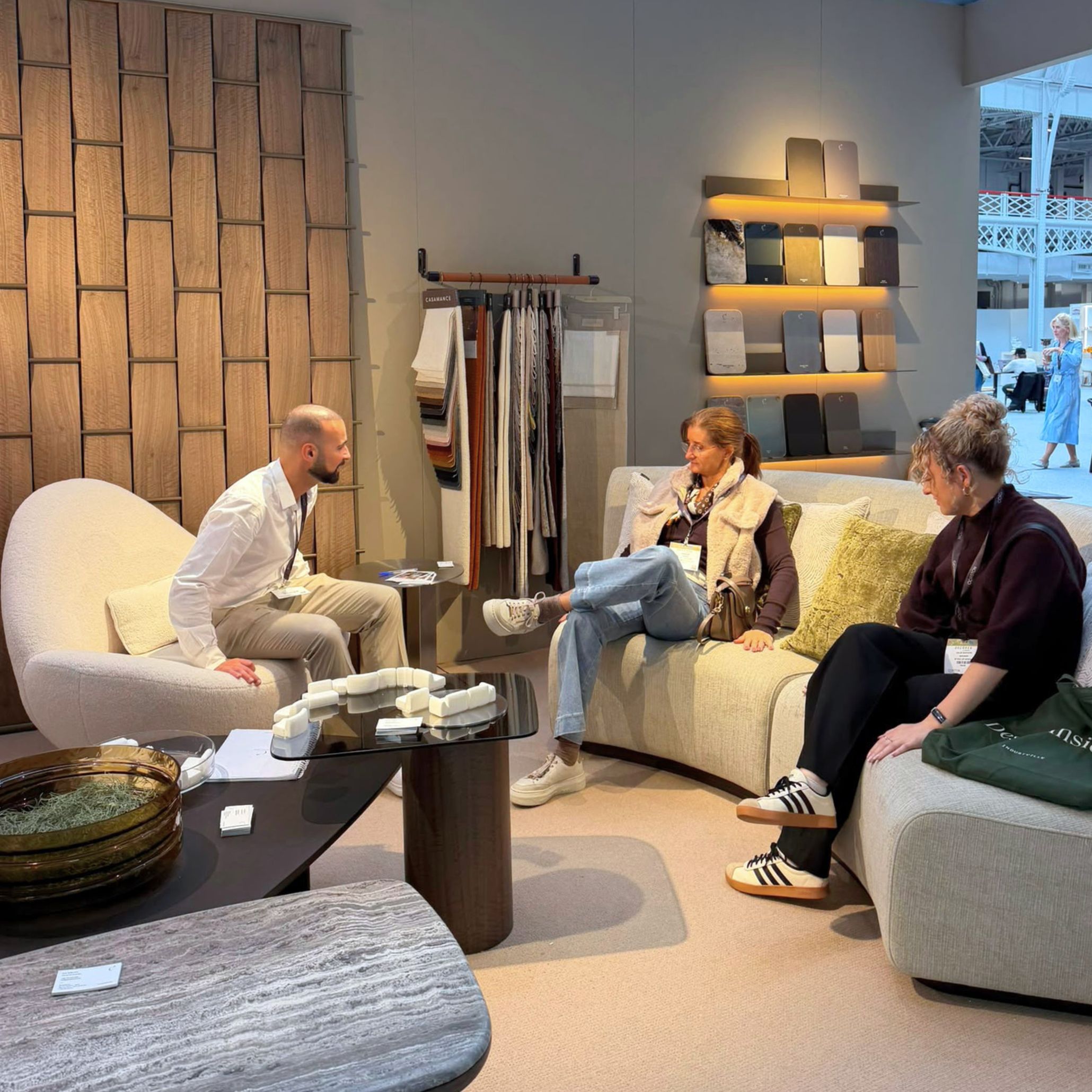 A representative conversing with interior designers seated on the modular Stone Sofa inside Stylish Club’s exhibition stand. Surrounded by material swatches and woven wood paneling, this space offers living room inspiration, high-end bespoke furniture solutions, and contemporary furniture luxury design.