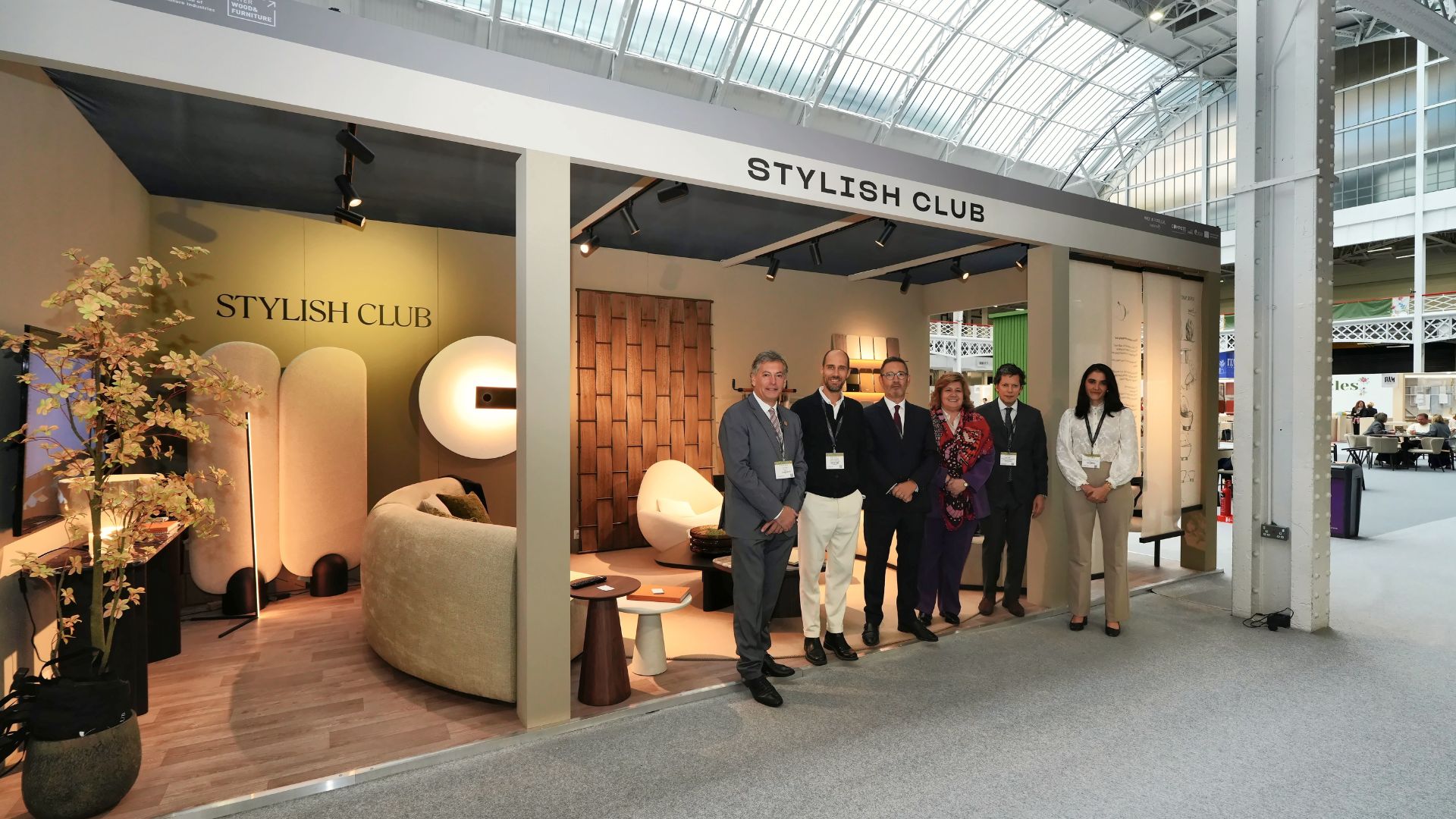 The Stylish Club team standing together in front of their exhibition stand at Decorex 2026 in London. The booth features organic curved seating, warm ambient wall lighting, and natural wood finishes, inviting visitors to discover interior design inspiration, high-end bespoke furniture solutions, and tailored custom furniture.