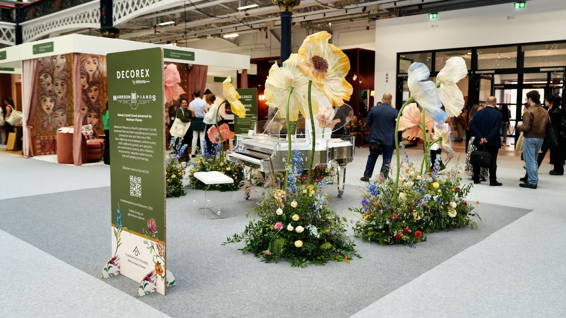 An artistic design installation at Decorex 2026 featuring a transparent grand piano surrounded by oversized floral sculptures and lush botanical arrangements. Visitors browse the exhibition floor, gathering interior design inspiration for modern spaces, showcasing how bespoke furniture and contemporary furniture can transform luxury interiors.