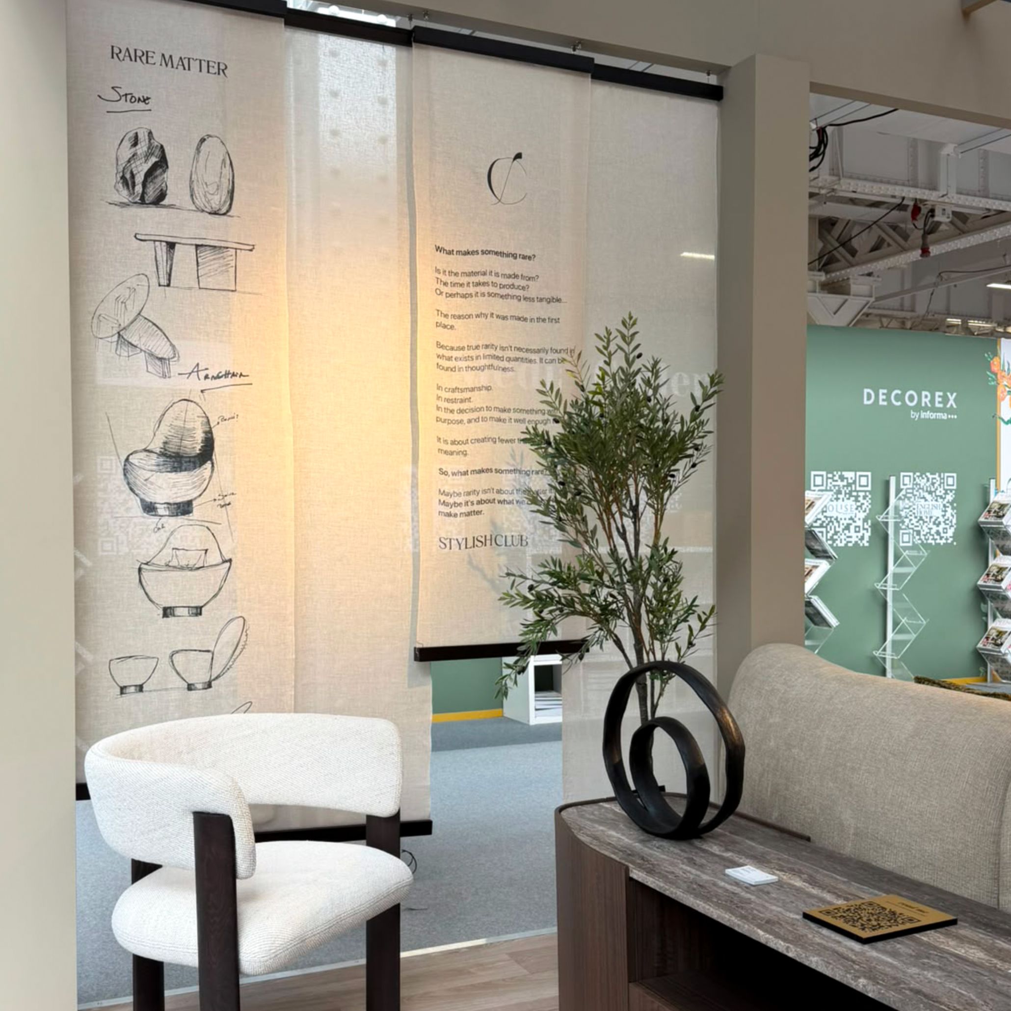 A serene wellness lounge corner inside Stylish Club’s stand at Decorex 2026, featuring a curved textured chair, dark wood finishes, and hanging linen banners with design sketches. The booth provides living room inspiration through tactile design details and custom furniture tailored for restorative, modern interiors.