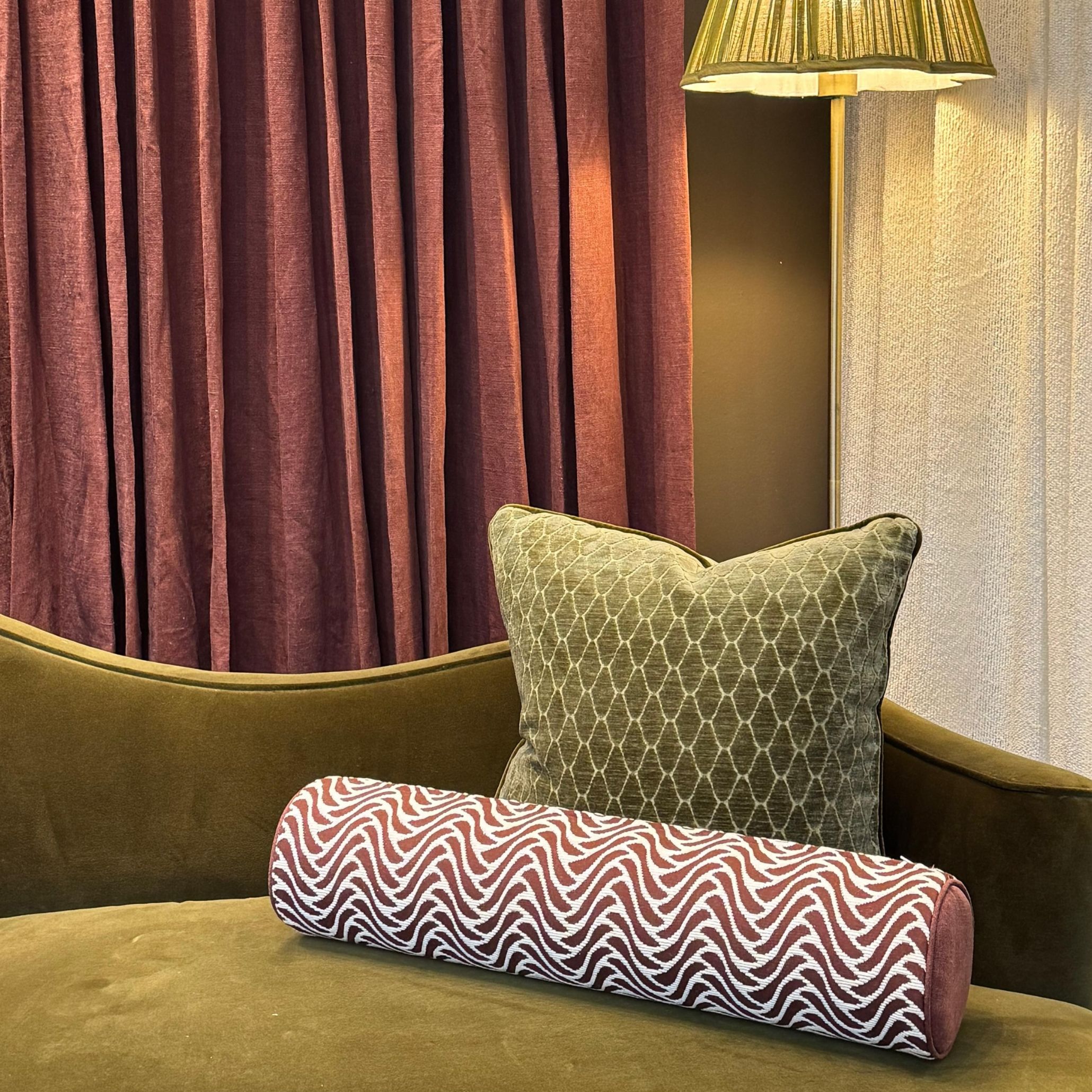 An elegant lounge seating arrangement featuring an olive green velvet daybed adorned with textured throw pillows against deep plum drapery. The rich color drenching and velvet furniture provide sophisticated living room inspiration using high-end furniture luxury design.