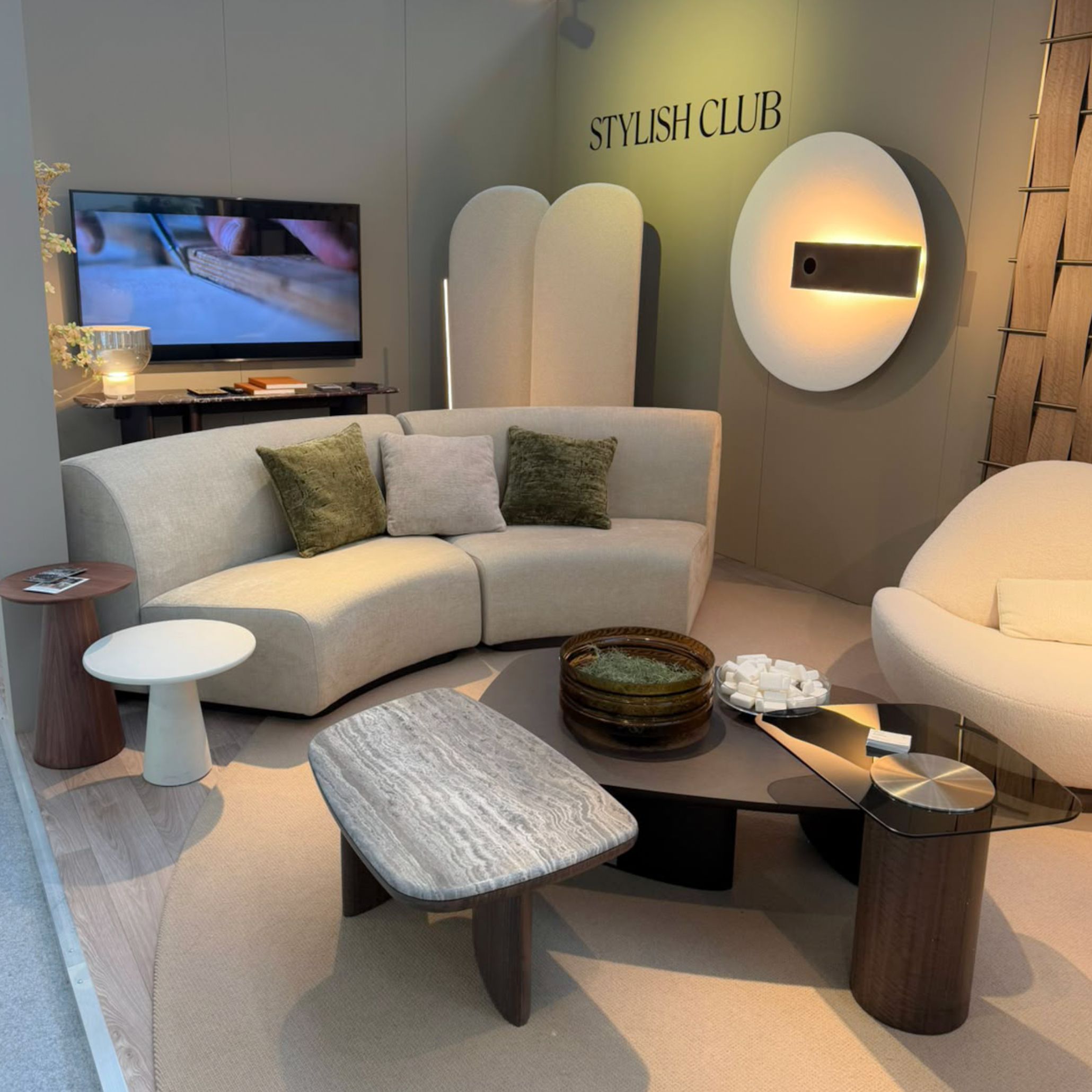 A modern living room layout at Stylish Club's Decorex 2026 stand showcasing the curved Stone Sofa, the Stone Armchair, and an expressive marble center table. The setup provides complete living room inspiration through organic silhouettes, artistic design details, and refined luxury living room furniture.