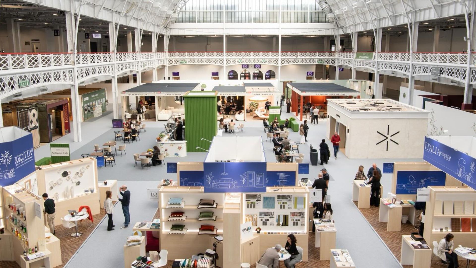 An elevated wide view of the Decorex 2026 exhibition hall, featuring illuminated design stands, curated display booths, and integrated lighting systems under a naturally lit glass roof atrium. The venue showcases modern interior design concepts that combine interior design inspiration, contemporary furniture, and modern luxury furniture into holistic wellness-driven environments.