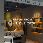 Stylish Club’s exhibition stand at Decorex 2026 featuring the title text "TRENDS FROM DECOREX 2026" overlaid across an warm interior display. The booth showcases plush modular seating, illuminated finish swatches, and organic wood paneling, offering sophisticated interior design inspiration, bespoke furniture solutions, and high-end custom furniture.
